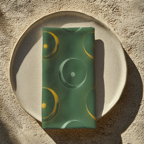 Four-piece cloth napkin set featuring a celestial print of radiant golden crescent moons and pale sunburst orbs against a rich emerald green background. Each napkin displays symmetrical, glowing motifs inspired by lunar phases and cosmic geometry. The fabric is neatly folded to showcase the repeating pattern, with a soft matte finish and fine detailing, perfect for mystical or magical table settings, altar decor, or celestial-inspired dining aesthetics.